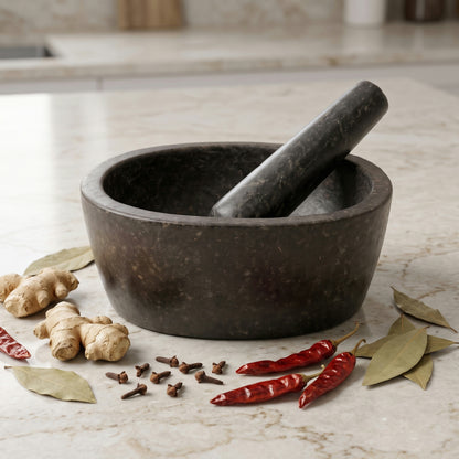 Oval granite mortar and pestle on a kitchen table with whole spices and fresh herbs beside it
